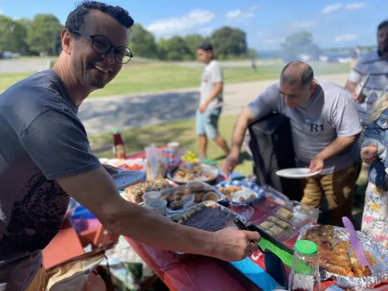 A smiling man serves himself food at an outdoor picnic table filled with dishes, while others help themselves in the background under a sunny sky.