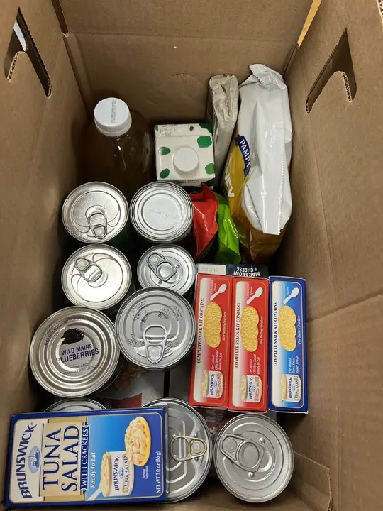 A cardboard box filled with canned food, a carton, a glass bottle, pasta packages, and a loaf of bread.
