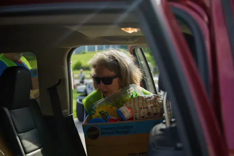 A woman wearing sunglasses loads a box of groceries into a car, seen through an open car door.