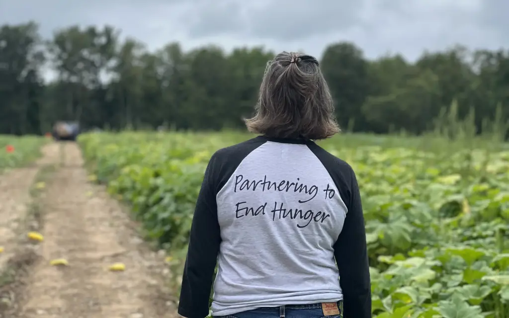 A person stands in a field, seen from behind, wearing a shirt that says Partnering to End Hunger. The sky is cloudy and there are green plants in rows.