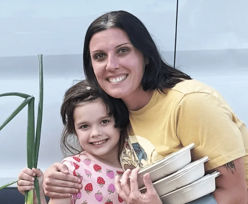 Smiling woman in yellow shirt hugs a young girl in a strawberry-print dress. The woman holds food containers; the girl holds a green plant. A light background is visible.