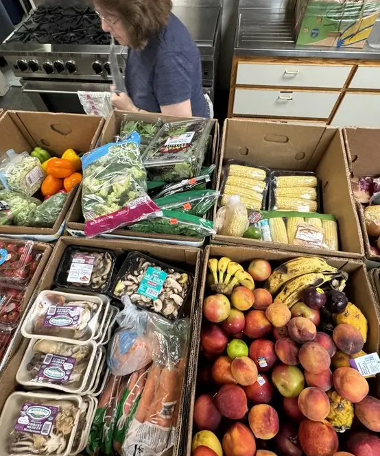 A woman stands behind boxes filled with fresh produce, including bananas, peaches, corn, mushrooms, bell peppers, and greens.